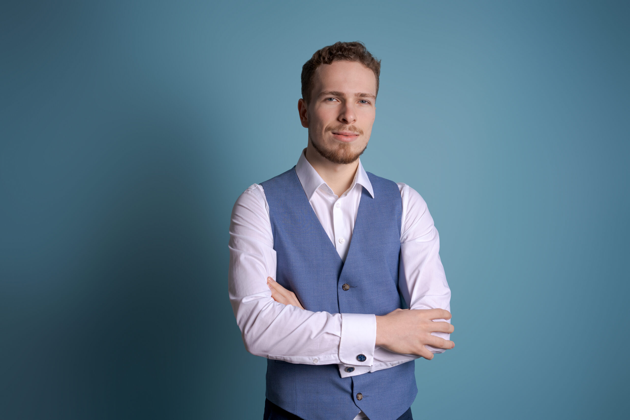 Young handsome man with beard in white shirt and blue vest standing on blue